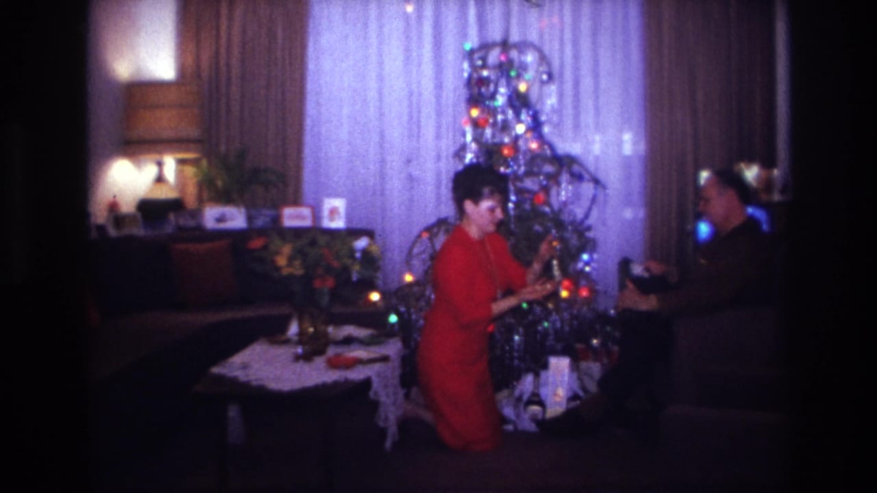 Woman reaches toward a Christmas tree as a man holds an item near the edge of the room.