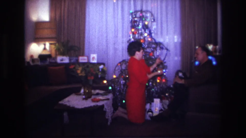 1967 Rockaway Beach home-movie frame showing a woman in a red dress kneeling near a tinsel Christmas tree with a seated man nearby.
