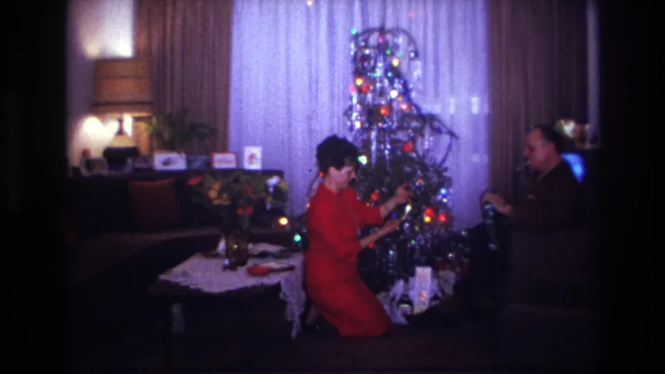 1967 Rockaway Beach home-movie frame of a woman adjusting a Christmas tree ornament in a private living room as a man sits nearby.