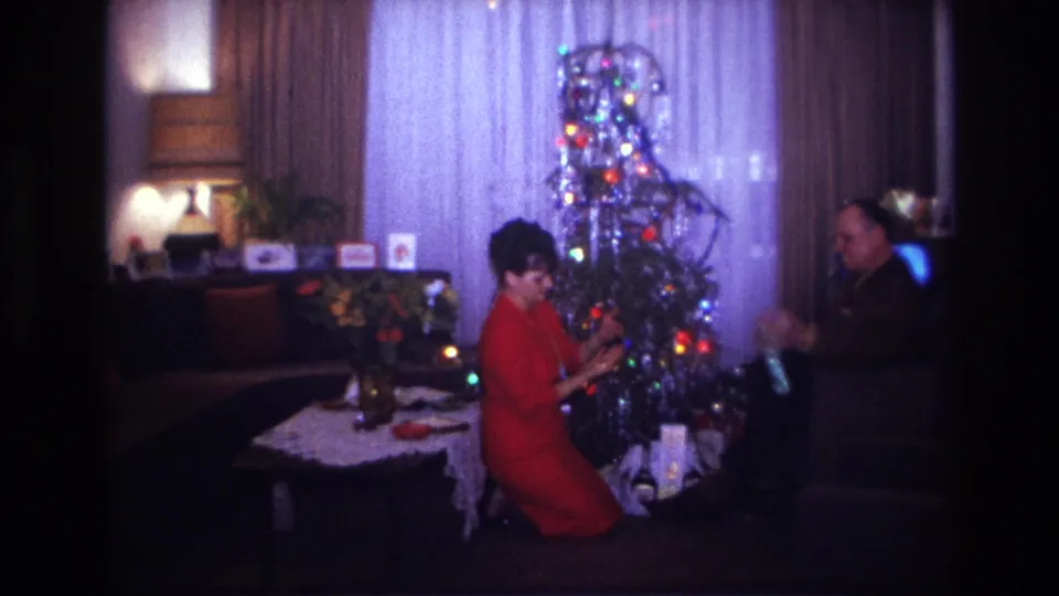 1967 Rockaway Beach home-movie frame showing a woman in a red dress by a silver Christmas tree as a seated man faces her.