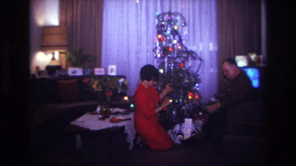 Home-movie frame from the 1967 Rockaway Beach holiday scene with two adults handling gifts beside a lit tinsel tree.