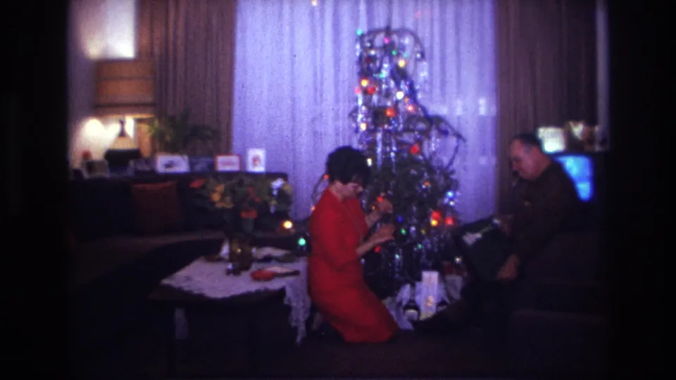 1967 home-movie frame showing a woman by a decorated tinsel tree while a seated man handles a dark gift box.