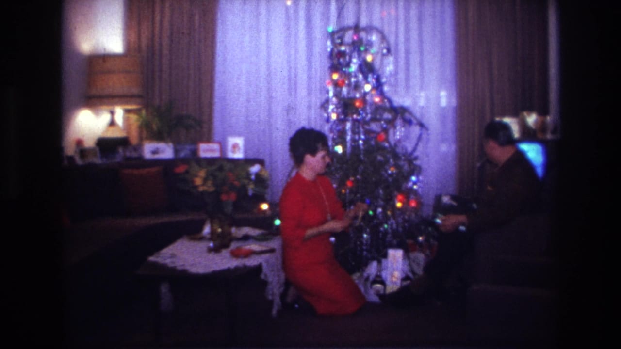 1967 Rockaway Beach home-movie frame showing a woman in red by a lit tinsel Christmas tree as a seated man handles gifts in a living room.