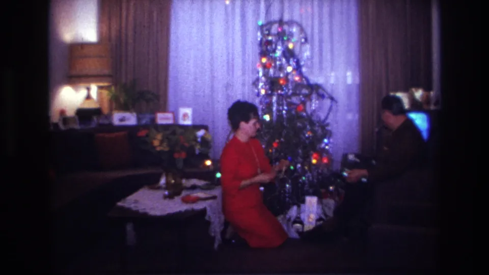 1967 Rockaway Beach home-movie frame showing a woman in red by a lit tinsel Christmas tree as a seated man handles gifts in a living room.