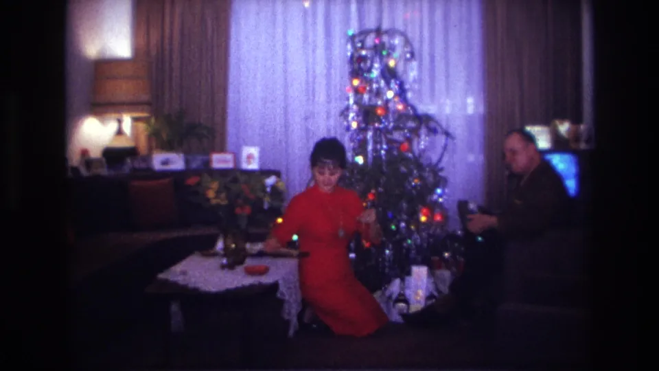 1967 Rockaway Beach living-room frame with a woman in red near the Christmas tree and a seated man beside gifts and a glowing television.