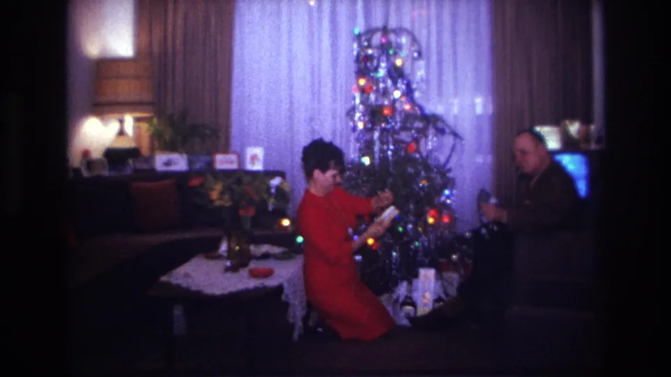 1967 Rockaway Beach home-movie frame showing a woman adjusting holiday decorations by a tinsel Christmas tree while a man sits close by.