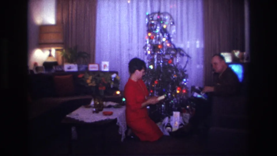 1967 Rockaway Beach frame with a woman in red examining an item by the lit Christmas tree as a seated man handles gifts nearby.