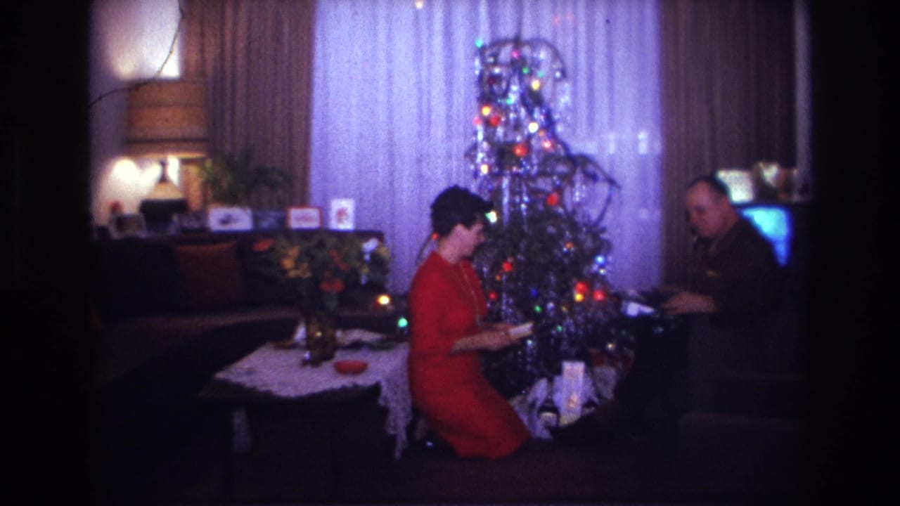 1967 Rockaway Beach home-movie frame showing a woman in a red dress beside a lit Christmas tree with a seated man nearby.
