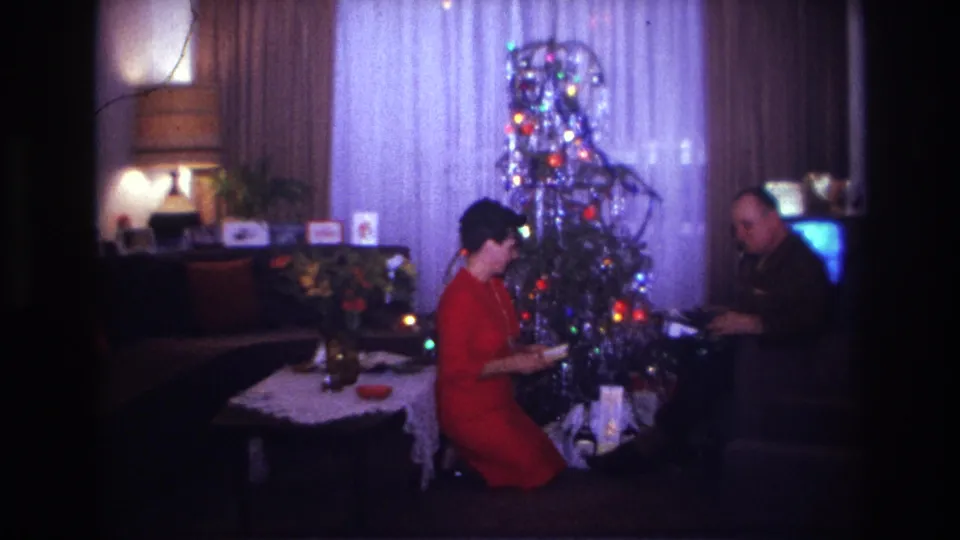 1967 Rockaway Beach home-movie frame showing a woman in a red dress beside a lit Christmas tree with a seated man nearby.