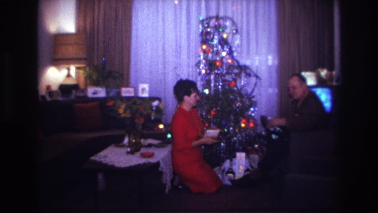 Woman and man sit near a lit Christmas tree with gifts in a living room.