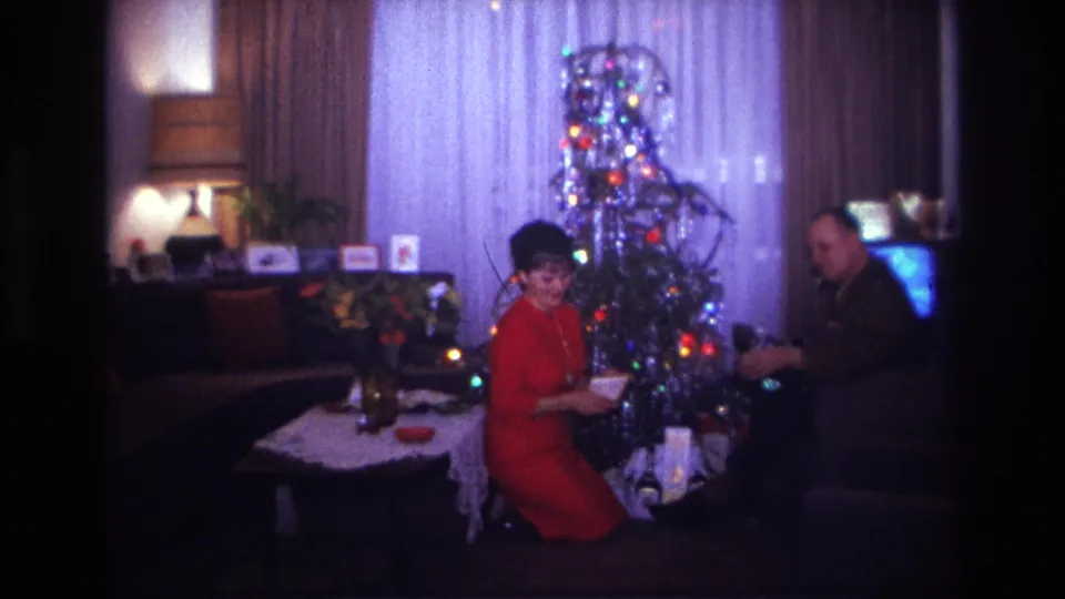 1967 home-movie frame of a woman in red looking toward the camera near a Christmas tree, presents, coffee table, and lamp.
