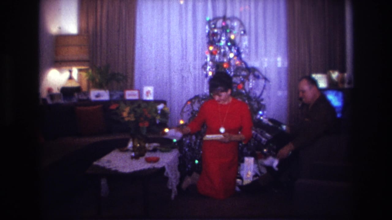 1967 Rockaway Beach home-movie frame of a woman in a red dress kneeling near a lit Christmas tree as a man sits beside her.