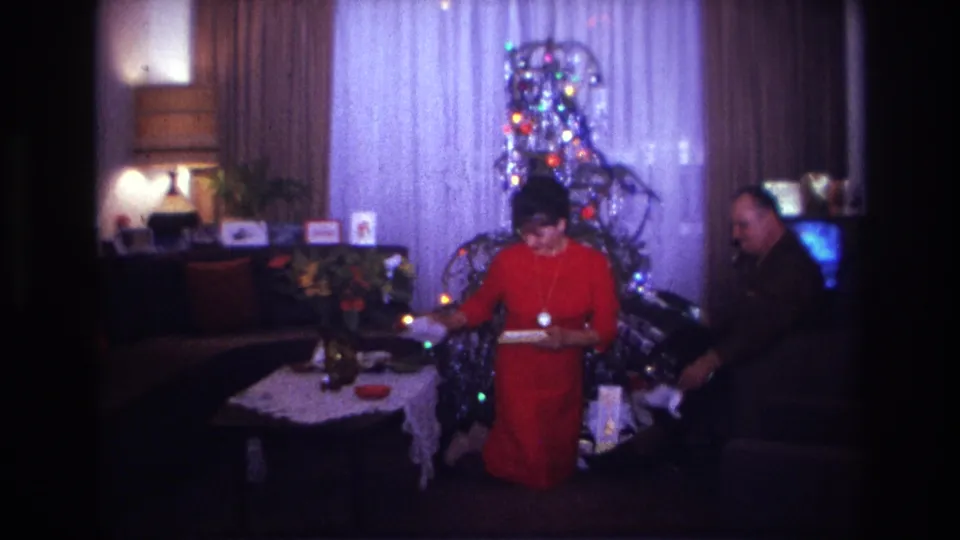 1967 Rockaway Beach home-movie frame of a woman in a red dress kneeling near a lit Christmas tree as a man sits beside her.