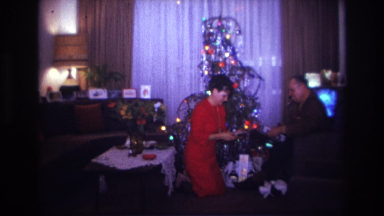 1967 home-movie frame from Rockaway Beach showing a kneeling woman and seated man exchanging or inspecting a gift beside the Christmas tree.