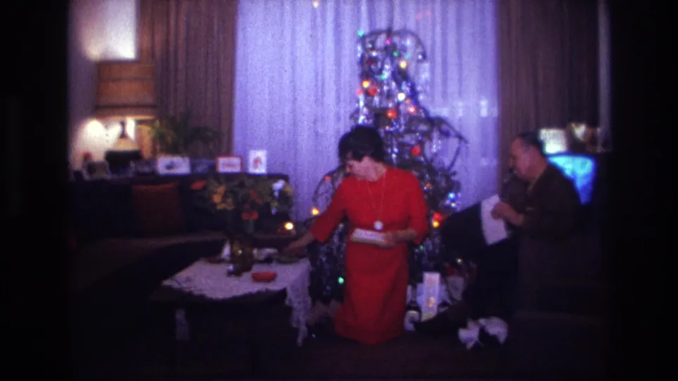 1967 Rockaway Beach living-room frame with a woman reaching toward a coffee table while a seated man holds paper near the Christmas tree.