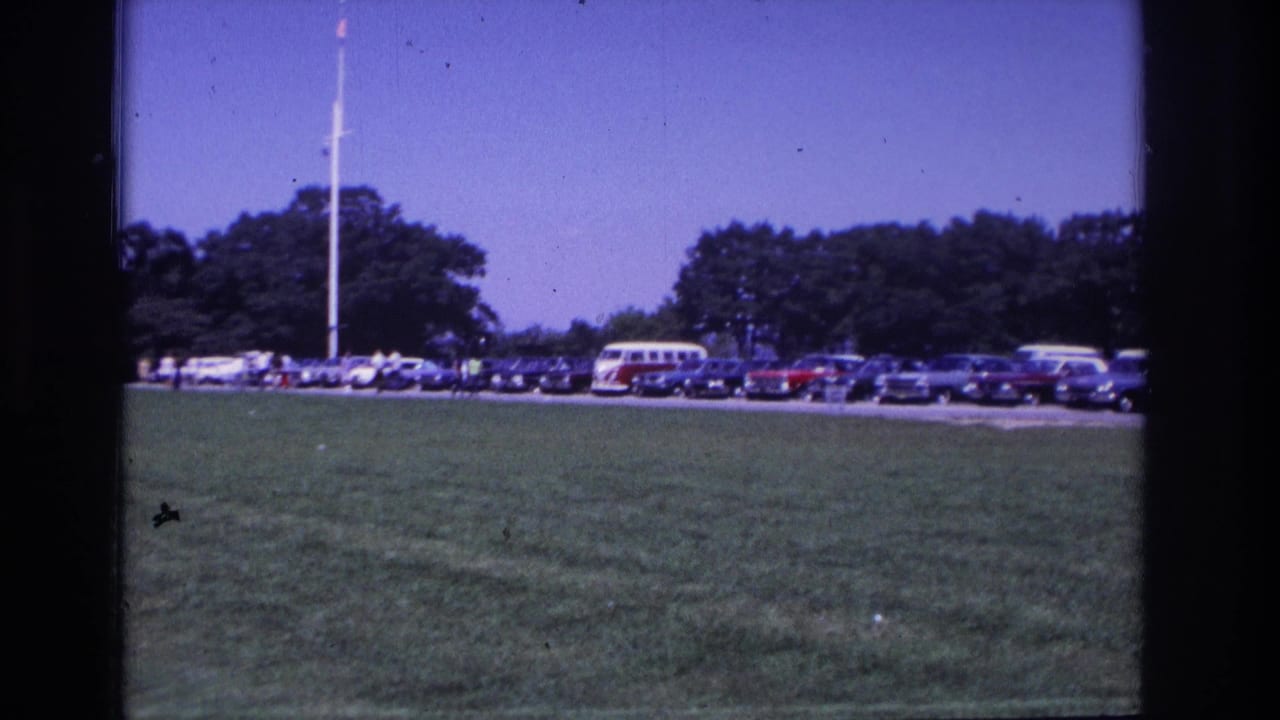 1968 Ocean City home-movie view of cars and a red-and-white VW bus parked beyond a grassy field near a flagpole and trees.