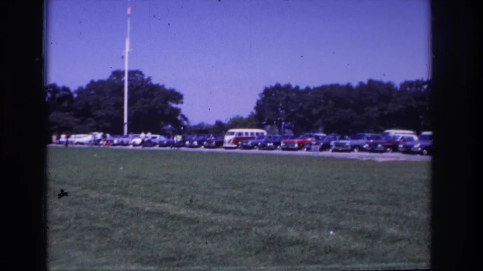1968 Ocean City home-movie view of cars and a red-and-white VW bus parked beyond a grassy field near a flagpole and trees.