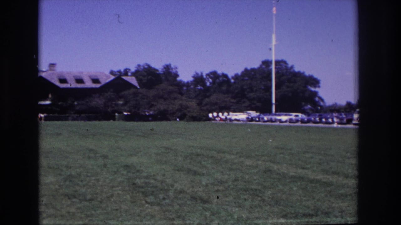 1968 Ocean City, New Jersey home-movie frame showing a grassy lawn, distant parked cars, trees, a flagpole, and a long dormered building.