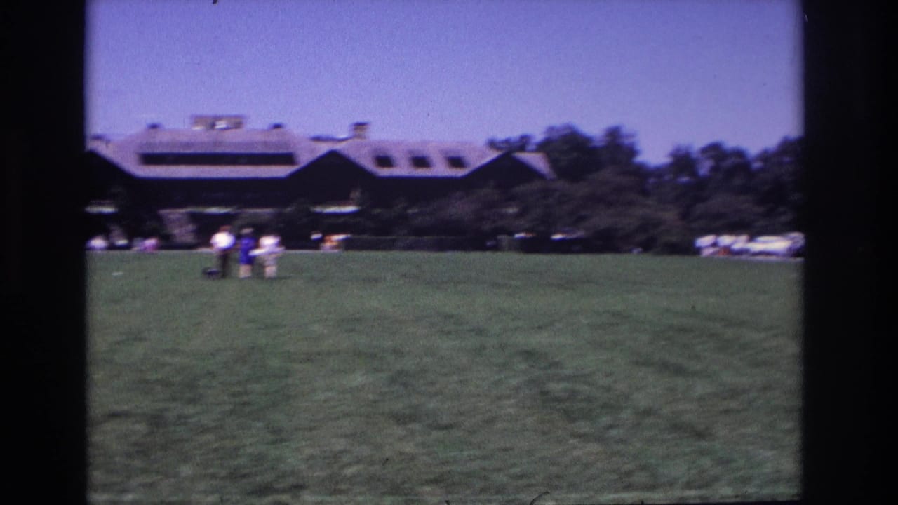 Small figures cross a grassy lawn in Ocean City, New Jersey, with a dark sloped-roof building and trees behind them.