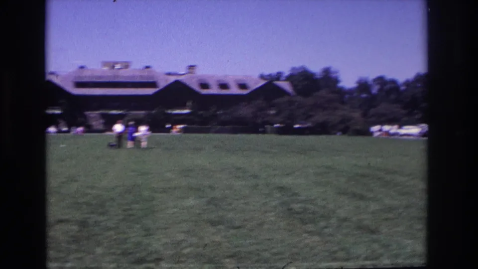 Small figures cross a grassy lawn in Ocean City, New Jersey, with a dark sloped-roof building and trees behind them.