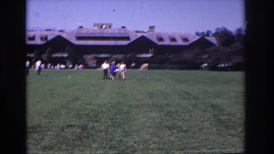 A 1968 home-movie view of people in 1960s attire on a lawn near a long building, with trees and open sky behind them.