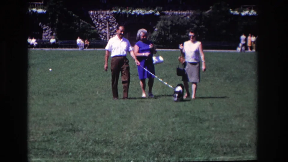 The 1968 home-movie frame shows three adults walking toward the camera on a park lawn while the small dog moves between them on a leash.