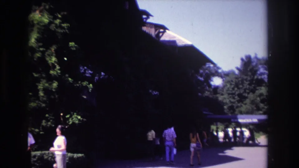 1968 Ocean City home-movie view of visitors walking along a paved path beside dense trees and a dark wooden structure.