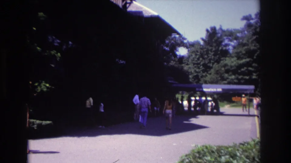 1968 Ocean City frame showing pedestrians on a sunny path beside a dark timber building and surrounding trees.