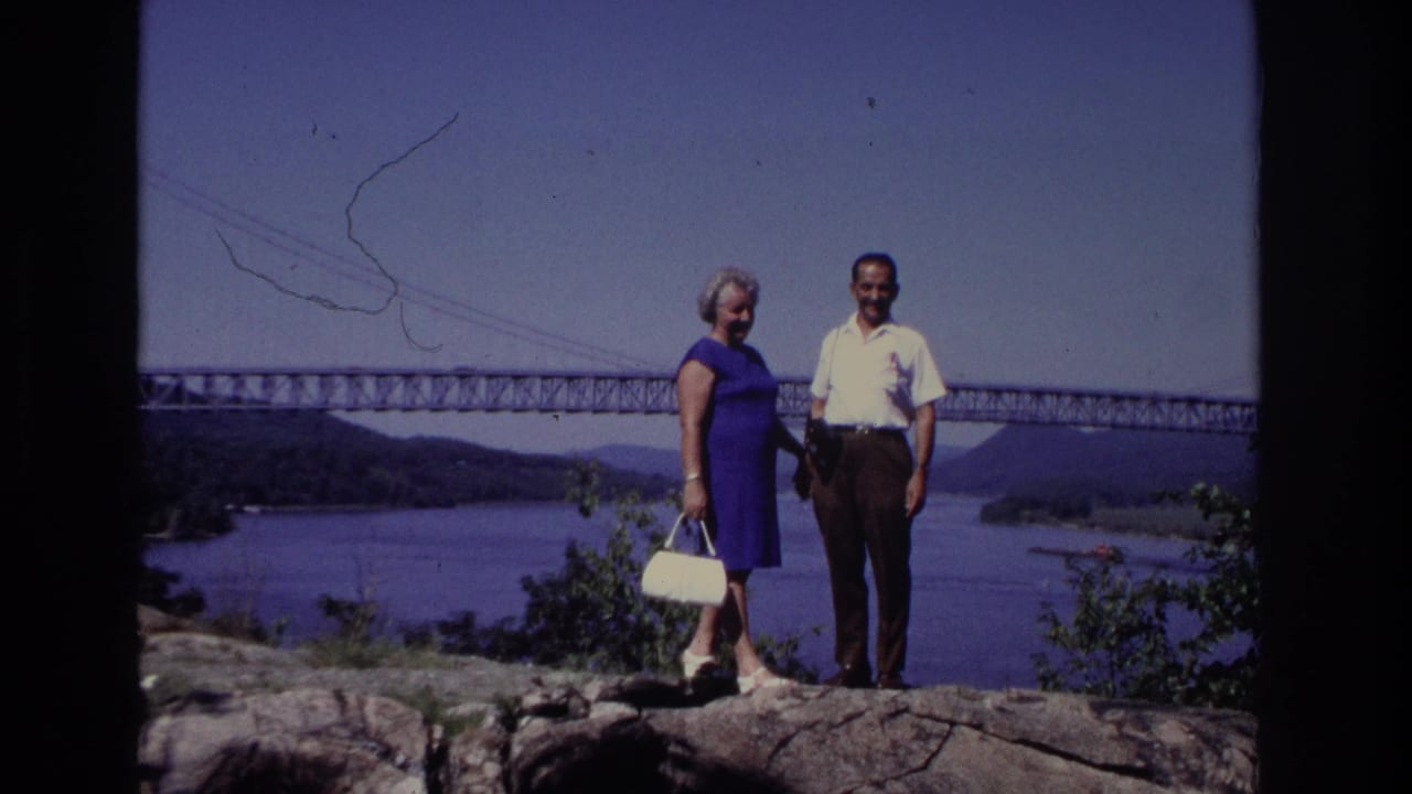 In the supplied 1968 Ocean City, New Jersey footage, a woman in a blue dress and a man in a white shirt pose on rocks above water with a long bridge behind them.