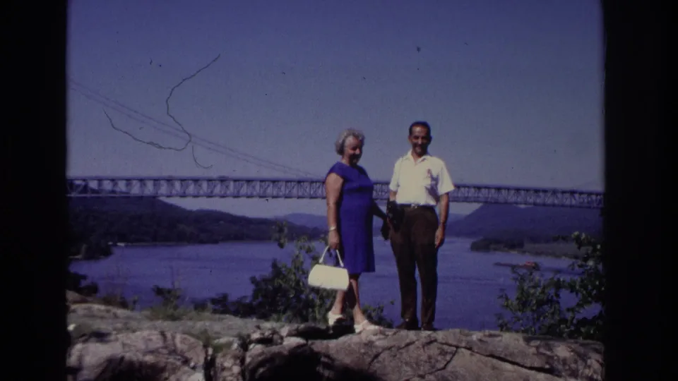 In the supplied 1968 Ocean City, New Jersey footage, a woman in a blue dress and a man in a white shirt pose on rocks above water with a long bridge behind them.