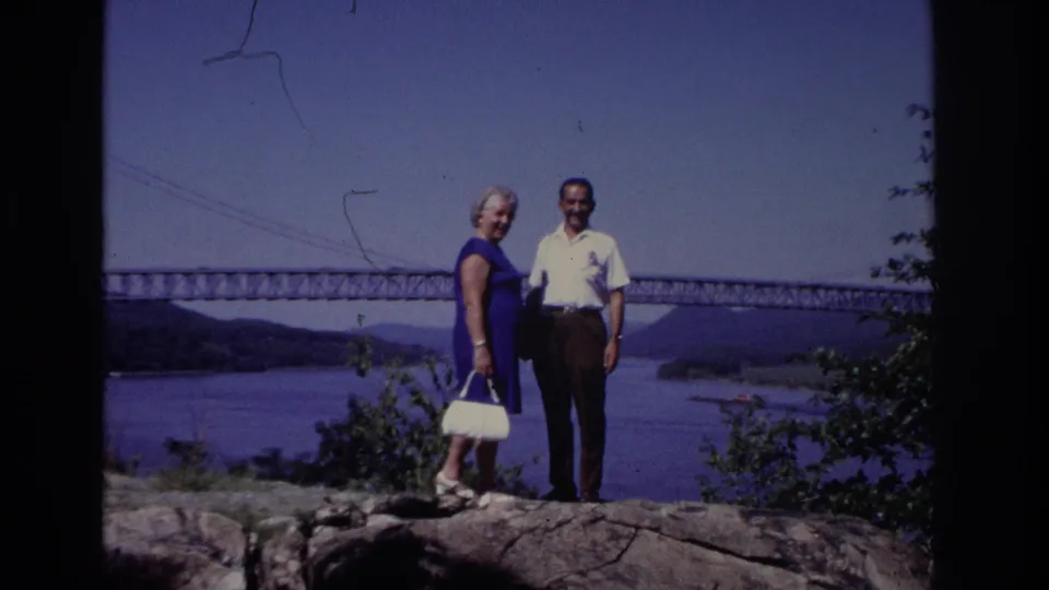 Final frame shows the smiling couple on the rocky overlook, with shrubs at the edge of the frame and a long bridge crossing the water behind them.