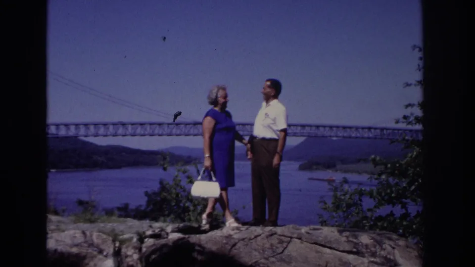 1968 8mm home-movie frame from Ocean City, New Jersey, showing two adults talking on a rocky overlook with a bridge and water behind them.