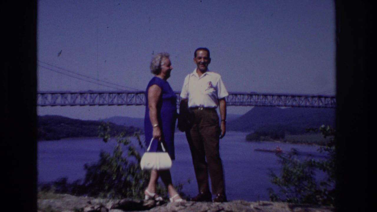 The couple stands closer to camera at the scenic overlook, with the woman holding a white handbag and the bridge crossing the water behind them.