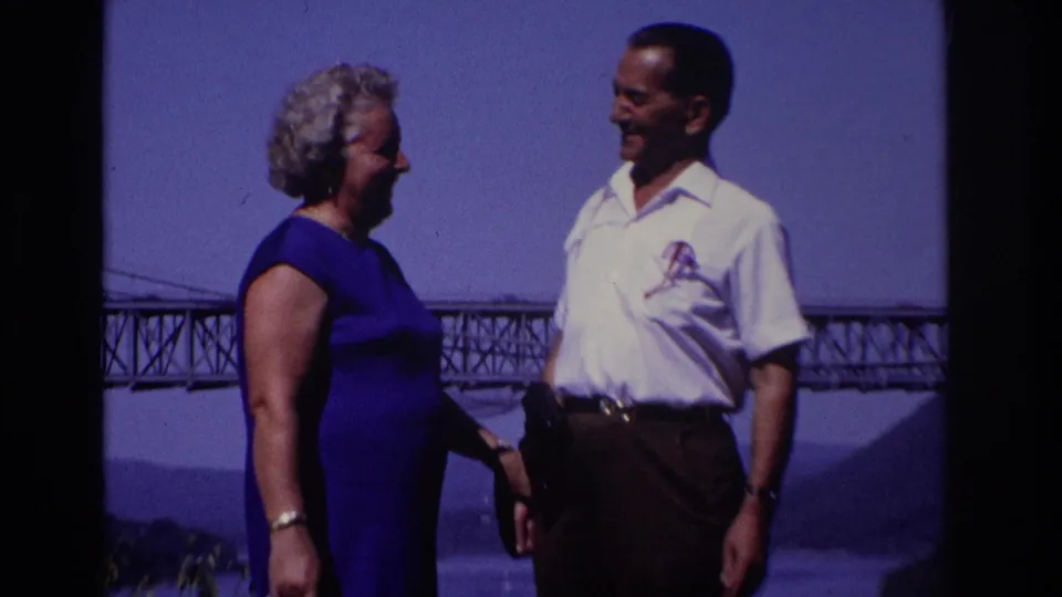 Closer 8mm frame of two adults smiling in conversation, with a truss bridge and water visible behind their shoulders.