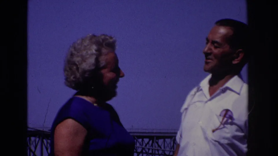 Tight side view of the adults in conversation, with blue sky, part of the bridge, and the man’s shirt emblem visible in the 1968 home-movie frame.