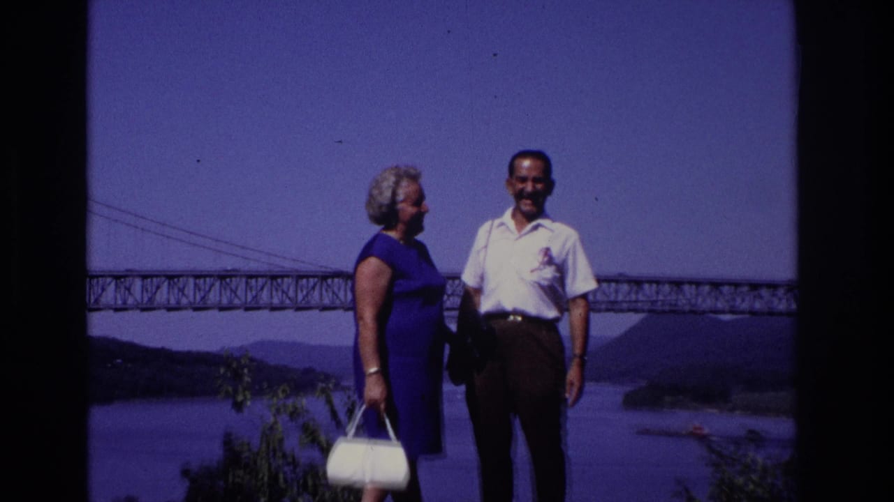 Adult couple pose on a rocky overlook in 1968 Ocean City, New Jersey, with a broad river and large bridge in the background.