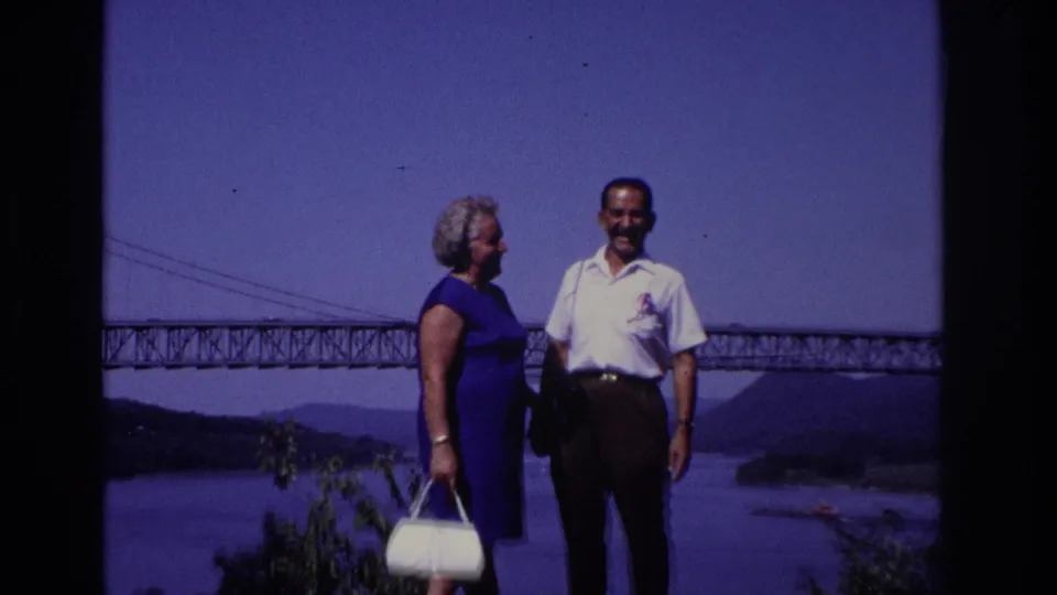 Adult couple pose on a rocky overlook in 1968 Ocean City, New Jersey, with a broad river and large bridge in the background.