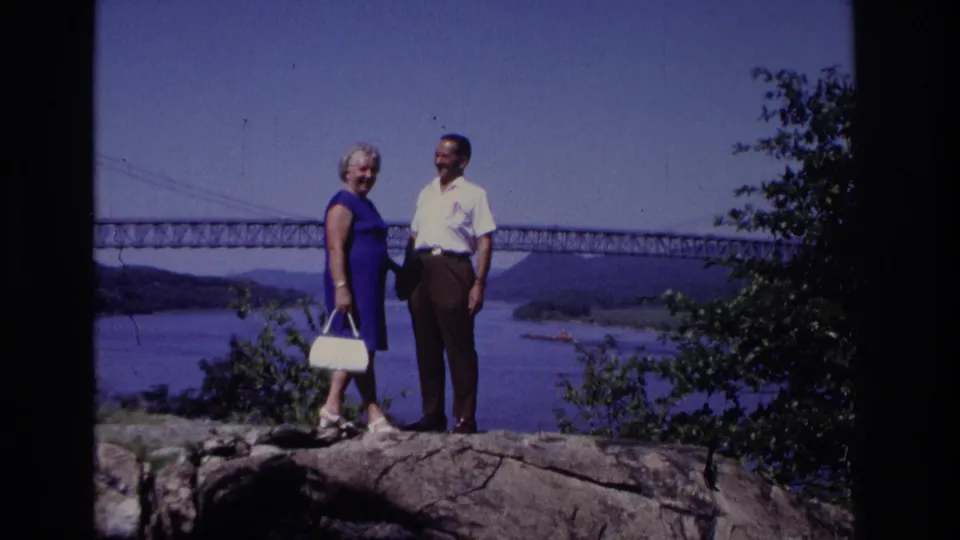 The woman in a blue dress holds a white handbag while standing with a man on rocks above the river and bridge.