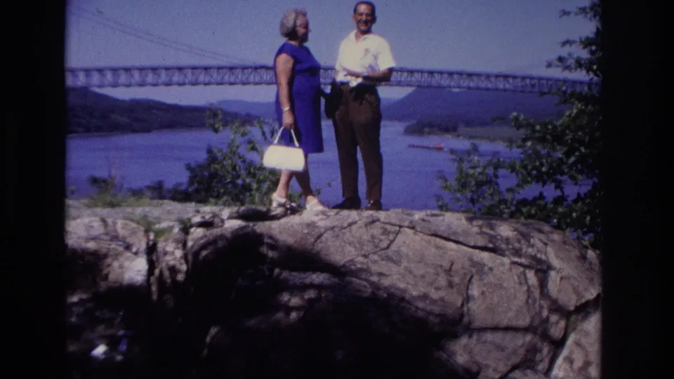 1968 home-movie frame of the couple standing on a rocky ledge beside shrubs, with a bridge and river landscape behind them.
