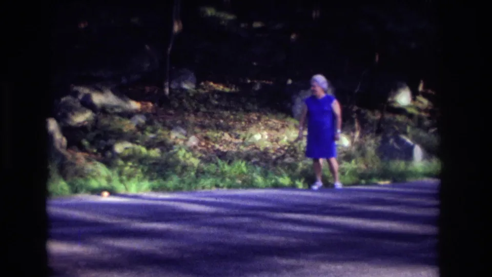 A woman in a blue dress stands on the road in the 1968 Ocean City, New Jersey home-movie scene, with rocks, grass, and trees behind her.