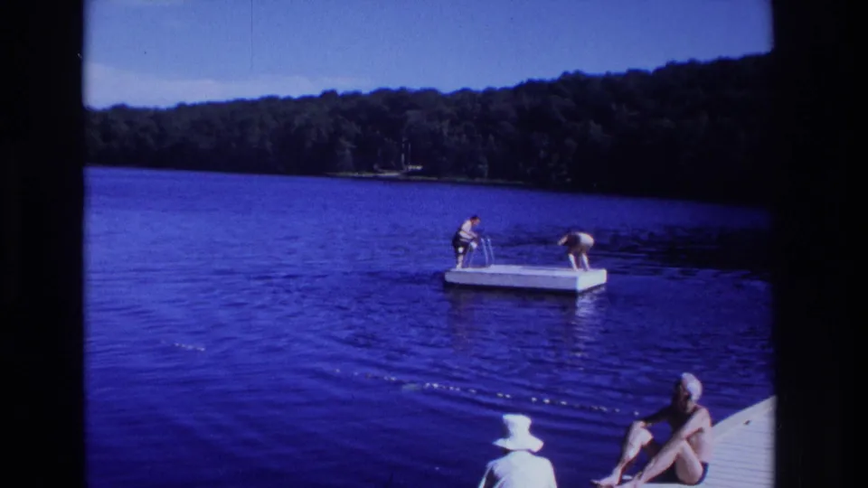 1968 home-movie frame showing people at a lake, two on a floating dock and another seated on a wooden pier, with wooded shoreline beyond.