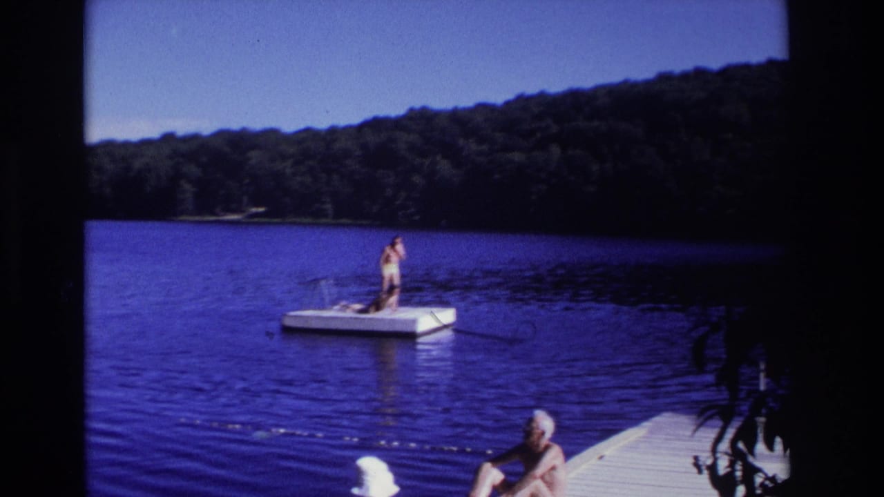 Person standing on a white floating dock in a blue lake, with an older man partly visible on a pier in the foreground.
