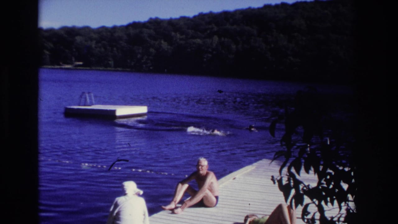 A seated man watches from a dock as swimmers move through the lake near a floating platform.