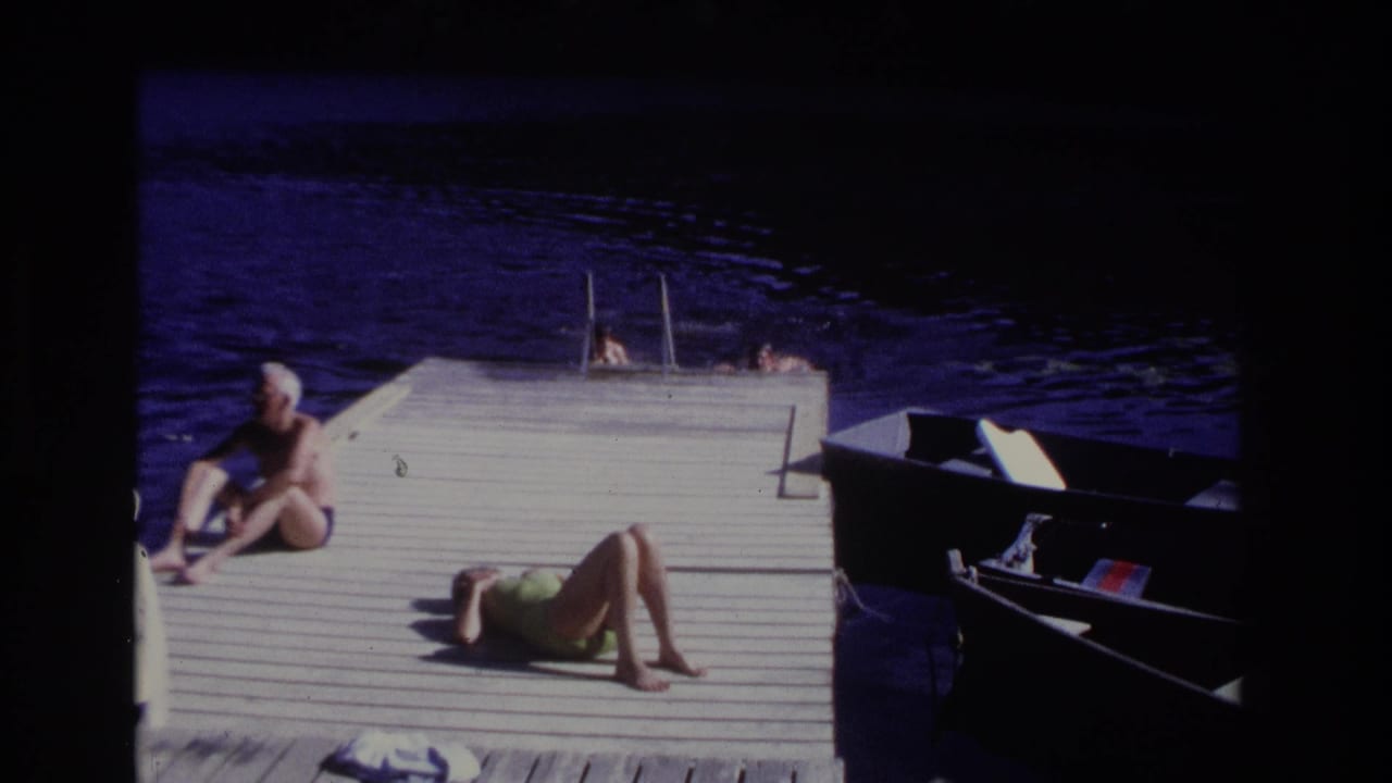 Adults sunbathe on a wooden dock while swimmers are in the water near a ladder.