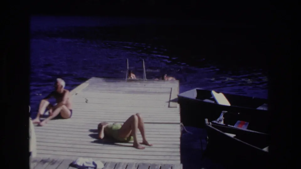 1968 Ocean City home-movie frame showing a seated man, a woman sunbathing in green, swimmers near a dock ladder, dark water, and a small boat.
