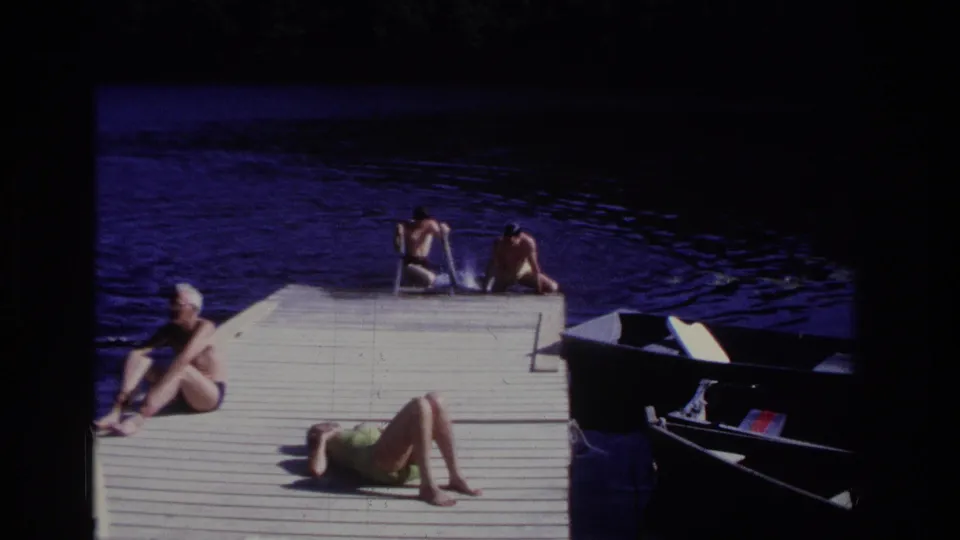 Ocean City 1968 home-movie frame with swimmers at the far edge of a wooden dock, adults sunbathing, dark water, and a small boat alongside.