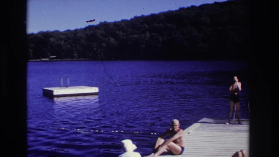 1968 Ocean City lakeside frame showing adults on a dock, a floating swim platform, blue water, and a tree-lined shoreline.