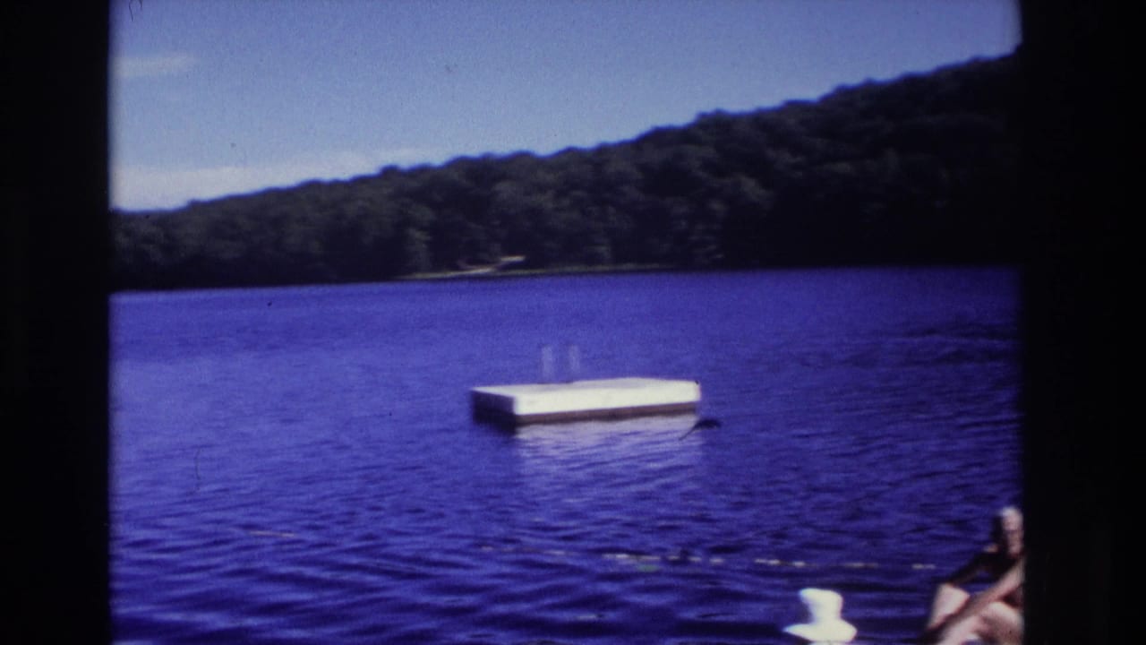 Blue lake with a floating swim platform, wooded shoreline, and a partial seated adult on a dock.