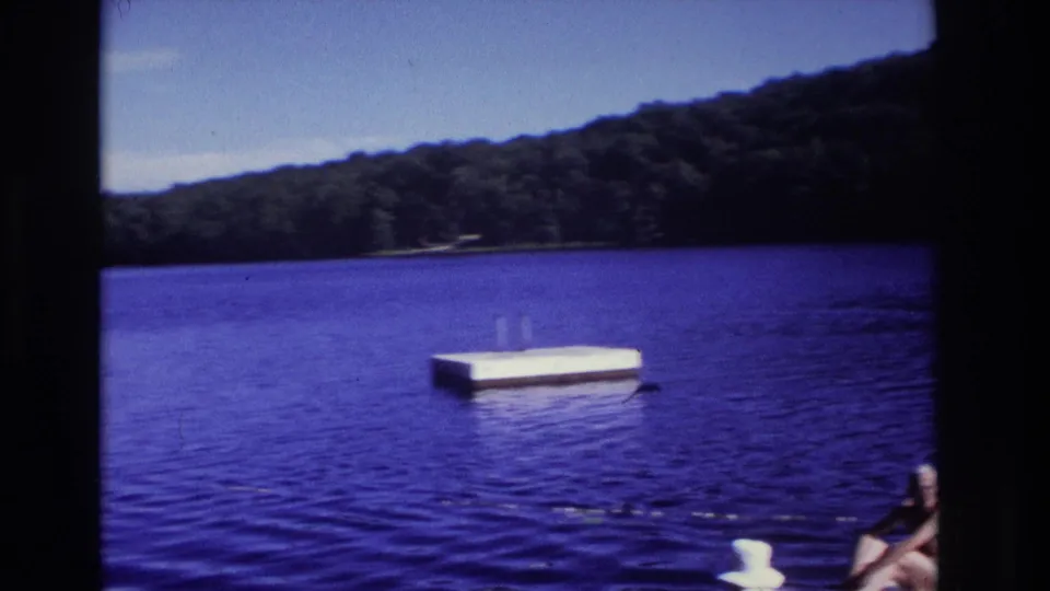1968 Ocean City, New Jersey home-movie frame showing a blue lake, floating platform, trees, dock edge, and partial seated adult figure.