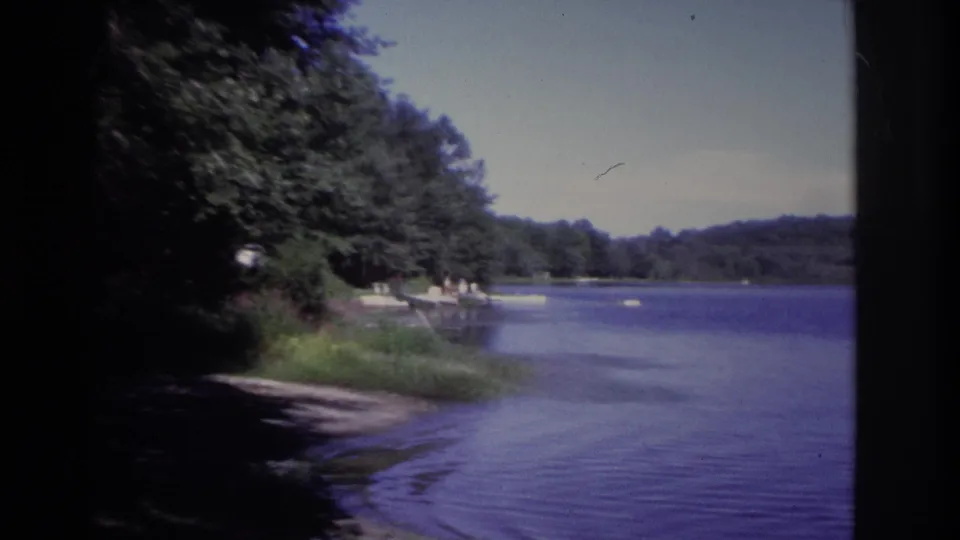 1968 Ocean City, New Jersey 8mm frame showing a shaded wooded shoreline, calm tinted lake water, small boats, and distant dock figures.